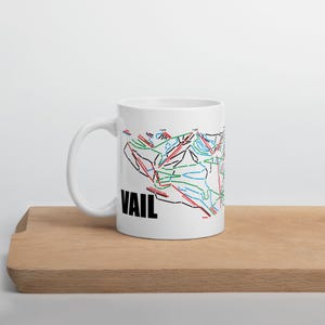 May include: White ceramic mug with a black and white illustration of the Vail ski resort map. The map is in black, red, blue, and green. The word "VAIL" is printed in black below the map.