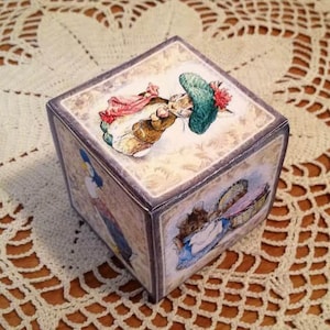 Puede incluir: Un cubo de madera con cuatro lados que presentan ilustraciones de personajes de Peter Rabbit. El cubo tiene un fondo marrón claro con un borde marrón oscuro. Los personajes están representados en un estilo vintage.