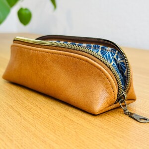 May include: A brown leather pencil case with a gold zipper and a blue and gold patterned interior. The case is rectangular with rounded edges and is partially open, revealing the patterned lining.