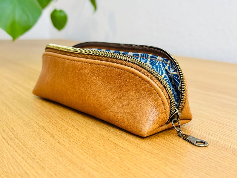 May include: A brown leather pencil case with a gold zipper and a blue and gold patterned interior. The case is rectangular with rounded edges and is partially open, revealing the patterned lining.