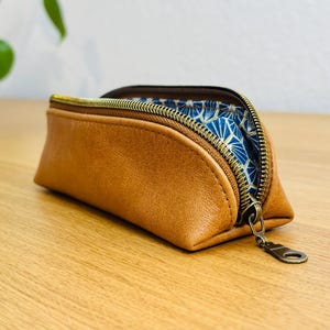 May include: A brown leather pencil case with a gold zipper and a blue patterned interior. The case is open, revealing the blue fabric. The zipper pull is also gold. The case is sitting on a wooden surface.