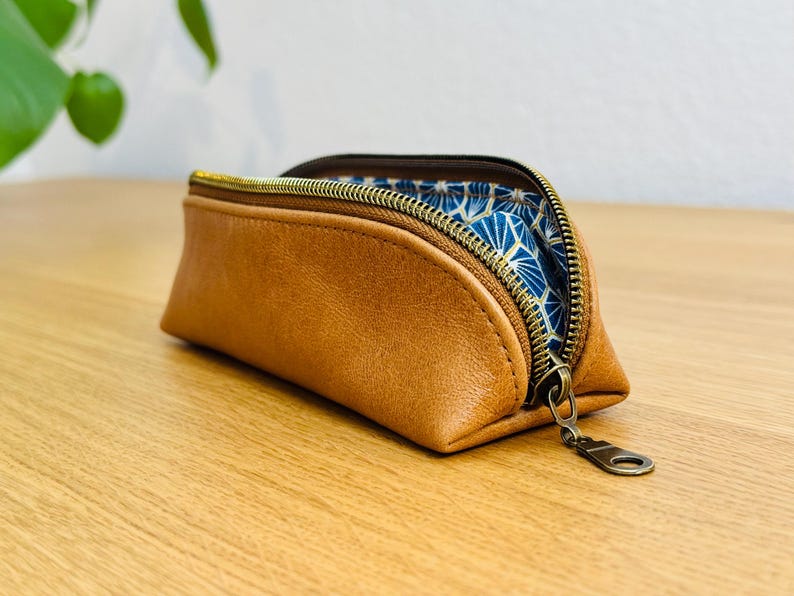 May include: A brown leather pencil case with a gold zipper and a blue patterned interior. The case is open, revealing the blue fabric. The zipper pull is also gold. The case is sitting on a wooden surface.