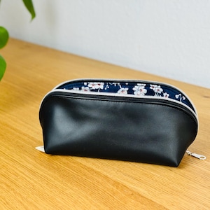 Könnte beinhalten: Schwarzes Federmäppchen aus Kunstleder mit weißem Reißverschluss und floral gemustertem Innenraum. Das Etui ist oval und liegt auf einer Holzoberfläche. Der Innenstoff hat einen marineblauen Hintergrund mit weißen und rosa Blumen.
