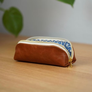 Può includere: Un astuccio in pelle marrone con finiture bianche e cerniera dorata. L'interno presenta un motivo floreale blu e bianco. L'astuccio è su una superficie di legno.