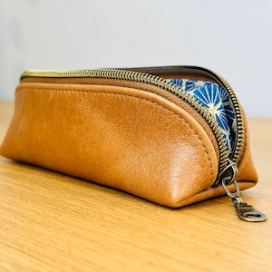 May include: A brown leather pencil case with a gold zipper. The interior lining is a blue fabric with a white and gold geometric pattern. The case is sitting on a wooden surface.