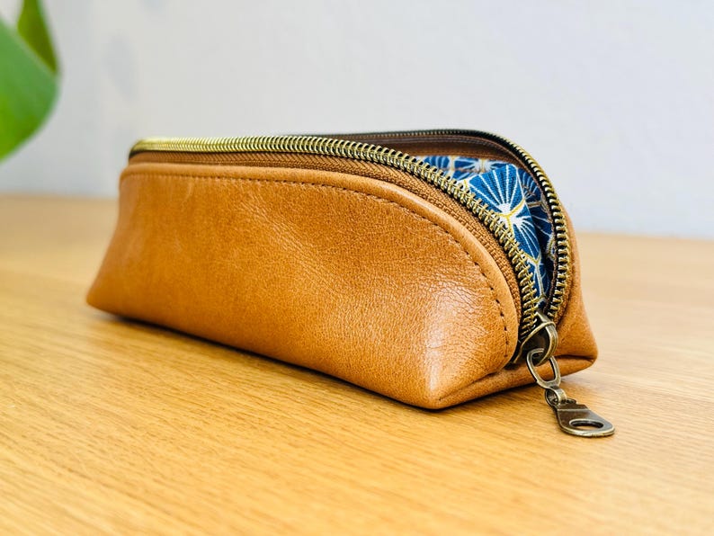 May include: A brown leather pencil case with a gold zipper. The interior lining is a blue fabric with a white and gold geometric pattern. The case is sitting on a wooden surface.