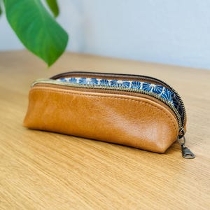 May include: A brown leather pencil case with a zipper closure. The interior lining features a blue and white floral pattern. The case is on a wooden surface.