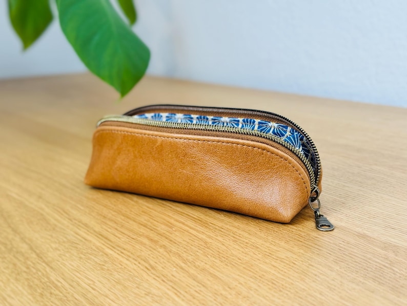 May include: A brown leather pencil case with a zipper closure. The interior lining features a blue and white floral pattern. The case is on a wooden surface.