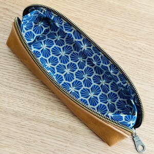May include: Open tan pencil case with a gold zipper and a blue geometric patterned interior. The pencil case is made of a leather-like material and is open, revealing the patterned lining.