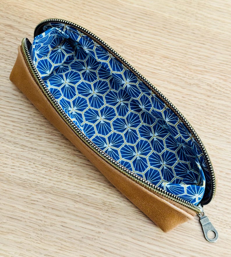 May include: Open tan pencil case with a gold zipper and a blue geometric patterned interior. The pencil case is made of a leather-like material and is open, revealing the patterned lining.