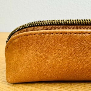 May include: Close-up of a tan leather pencil case with a brass zipper. The case has a rounded shape and visible stitching along the seams. The leather has a textured appearance.