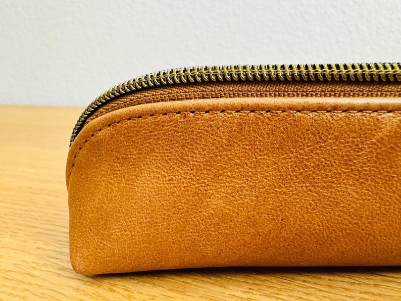 May include: Close-up of a tan leather pencil case with a brass zipper. The case has a rounded shape and visible stitching along the seams. The leather has a textured appearance.