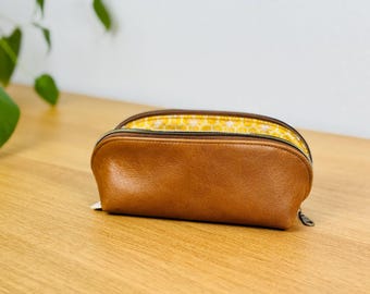 May include: A brown, semi-oval cosmetic bag with a zipper closure. The bag's exterior is a smooth, textured brown, and the interior lining features a yellow and white patterned fabric. The bag is open, revealing the interior.
