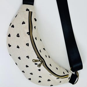 Puede incluir: Riñonera beige con estampado de corazones negros. La bolsa tiene una correa negra ajustable y una cremallera dorada. Diseñada para llevarse cruzada o en la cintura. Fabricada con tejido resistente.