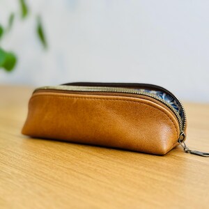 May include: A brown leather pencil case with a zipper closure. The case has a rounded shape and a patterned interior. The zipper is a dark metal color. The case is sitting on a wooden surface.