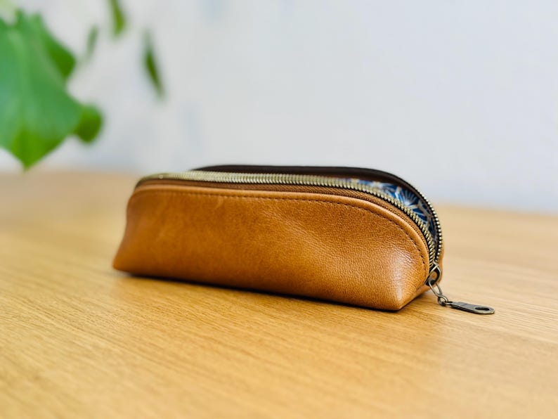 May include: A brown leather pencil case with a zipper closure. The case has a rounded shape and a patterned interior. The zipper is a dark metal color. The case is sitting on a wooden surface.