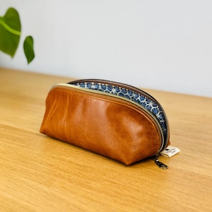 Könnte beinhalten: Ein braunes Leder-Federmäppchen mit goldenem Reißverschluss und blau-weiß gemustertem Innenfutter. Das Etui ist oval und liegt auf einer Holzoberfläche. Ein kleines Etikett ist am Reißverschluss befestigt.