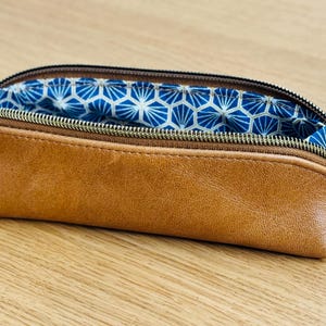 May include: A brown leather pencil case with a zipper closure. The interior lining features a blue and white geometric pattern. The pencil case is open, revealing the patterned lining. The zipper pull is a dark metal color.