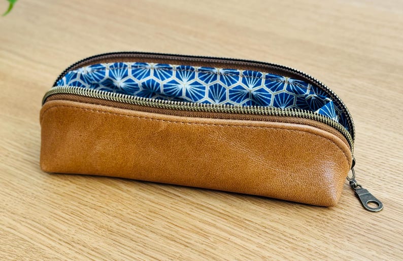 May include: A brown leather pencil case with a zipper closure. The interior lining features a blue and white geometric pattern. The pencil case is open, revealing the patterned lining. The zipper pull is a dark metal color.