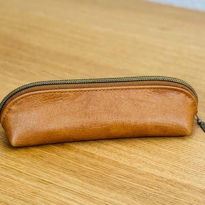 May include: A brown leather pencil case with a bronze zipper. The rectangular case is made of smooth, textured leather. The zipper pull is a small, round metal loop. The case is on a light brown wooden surface.