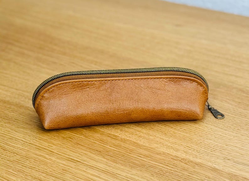 May include: A brown leather pencil case with a bronze zipper. The rectangular case is made of smooth, textured leather. The zipper pull is a small, round metal loop. The case is on a light brown wooden surface.