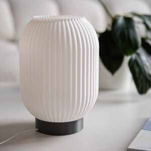 May include: White table lamp with a ribbed, cylindrical shade and a dark gray base. The lamp is sitting on a white table with a stack of books.
