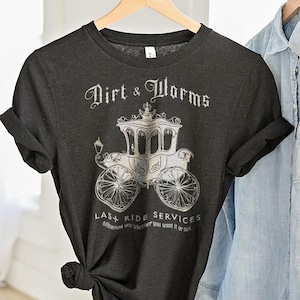 May include: Dark gray t-shirt with the text "Dirt & Worms" above a vintage carriage illustration. The shirt also features the text "Last Ride Services". The shirt is hanging on a wooden hanger.