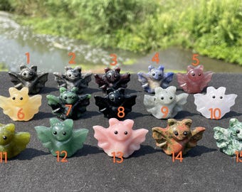 Puede incluir: Una colección de quince figuras de murciélagos de piedra tallada en varios colores, incluyendo negro, verde, rosa y amarillo. Los murciélagos se exhiben sobre una superficie oscura con un fondo natural.
