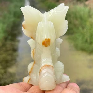May include: A carved onyx sculpture of a stylized bird or mythical creature. The sculpture is primarily off-white with streaks of brown and orange. The wings are spread, and the overall design is symmetrical. The sculpture is held in a hand.