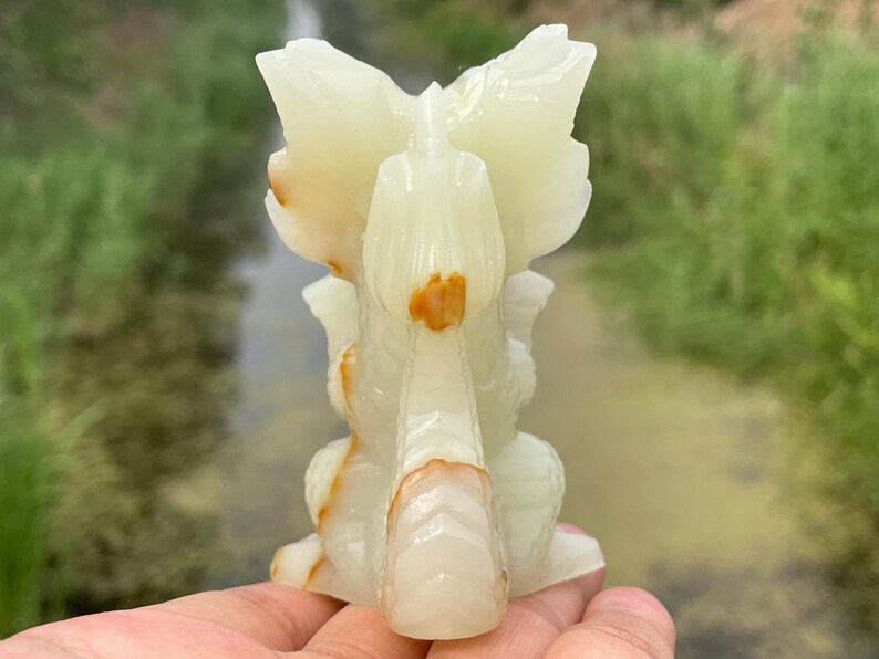 May include: A carved onyx sculpture of a stylized bird or mythical creature. The sculpture is primarily off-white with streaks of brown and orange. The wings are spread, and the overall design is symmetrical. The sculpture is held in a hand.
