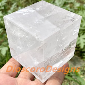 Pode incluir: Um grande cubo de cristal transparente com uma superfície lisa e polida. O cubo tem aproximadamente 7,5 cm de lado.