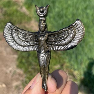 Op de afbeelding: Een zilverkleurige metalen hanger met een afbeelding van een gevleugelde godin met een kroon. De hanger is gedetailleerd met ingewikkelde gravures en heeft een licht verweerde afwerking.
