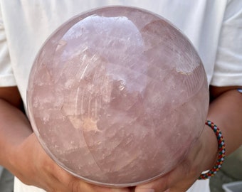 May include: A large, polished rose quartz sphere. The sphere is a soft pink color with natural variations in the stone. The smooth surface reflects light, creating a subtle glow. The sphere is held in both hands.
