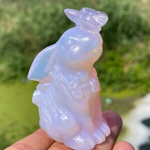 May include: A translucent, opalescent bunny figurine with a hat. The bunny is sitting upright, with its ears perked up. The figurine has a smooth, polished surface and is held in a hand against a blurred green background.