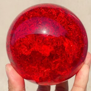 Peut inclure: Une grande boule de cristal rouge et ronde avec un intérieur texturé et scintillant. La sphère est tenue dans une main, mettant en valeur sa surface polie et réfléchissante. La boule de cristal est un objet décoratif.