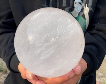 May include: A large, clear quartz crystal sphere. The sphere is held in front of a person, and it has a smooth, polished surface. The crystal ball is a translucent white color and appears to be made of natural stone.