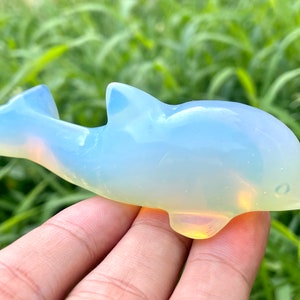 May include: A white opal dolphin figurine. The dolphin is carved from a translucent white opal with a slight yellow hue. The figurine is approximately 7.5 cm long and 2.5 cm wide.