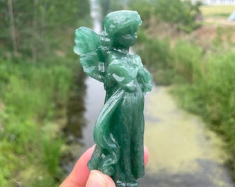 May include: A green jade angel figurine with detailed wings and flowing robes. The statue is carved from a translucent, polished stone, showcasing the natural variations in the jade. The angel is depicted in a graceful pose, with a serene expression.
