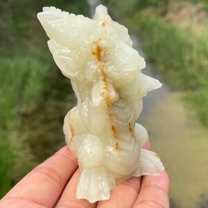 May include: A carved, off-white onyx sculpture of a bird-like creature with detailed feathers and a textured surface. The sculpture has brown and orange veining throughout. The sculpture is held in a hand.