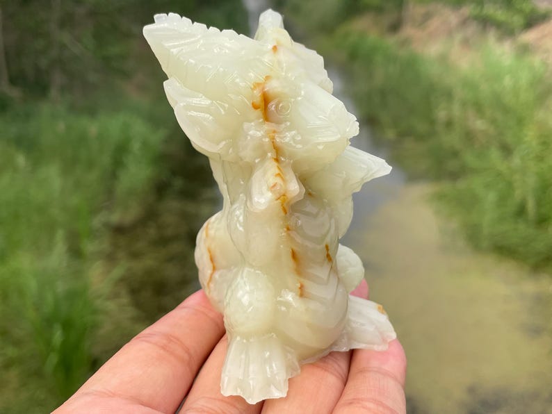 May include: A carved, off-white onyx sculpture of a bird-like creature with detailed feathers and a textured surface. The sculpture has brown and orange veining throughout. The sculpture is held in a hand.