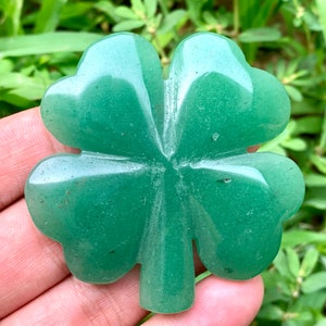 Pode incluir: Um trevo de quatro folhas esculpido em aventurina verde. O trevo é liso e polido, com uma cor verde clara.