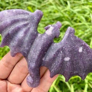 May include: A purple lepidolite crystal carved into the shape of a bat. The crystal has a sparkly, iridescent finish.