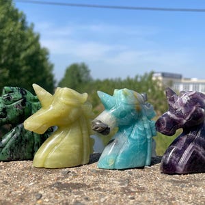 May include: Four carved stone unicorn figurines in shades of green, yellow, blue, and purple. The unicorns are standing on a rough, textured surface, with a backdrop of trees and a blue sky.