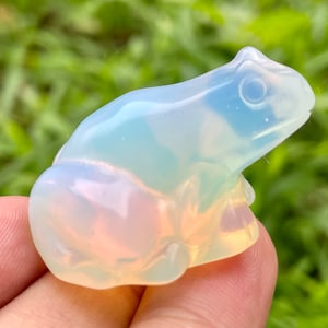 Puede incluir: Una pequeña figura de rana de ópalo blanco con colores arcoíris iridiscentes. La rana está sentada en una mano con un fondo verde borroso.