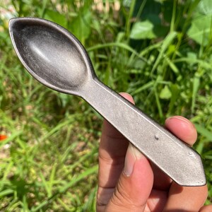 Puede incluir: Una cuchara gris oscuro con un mango largo y un cuenco ovalado. La cuchara se sostiene contra un telón de fondo de hierba verde y follaje. La cuchara parece ser de metal.