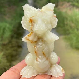 May include: A carved, off-white onyx figurine of a creature with a stylized head, wings, and a rounded body. The figurine has brown and orange veining throughout. The sculpture is held in a hand against a blurred green background.