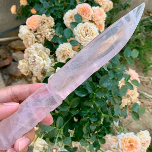 May include: A pink crystal knife with a clear blade and a textured handle. The knife is held in a hand against a background of pink and white roses.