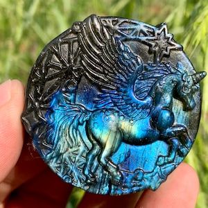 May include: A round, blue and green labradorite gemstone pendant carved with a winged unicorn. The unicorn is running with its wings spread out. The pendant is set in a silver tone metal.