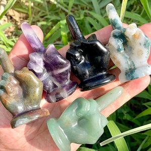 May include: Five small hand sculptures carved from different types of stone. Each hand is making the middle finger gesture. The stones are green, black, purple, and white with green and brown markings.