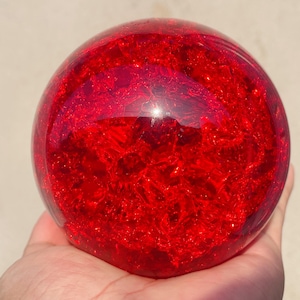 Puede incluir: Una esfera grande y redonda de vidrio rojo con una apariencia agrietada y texturizada. La esfera se sostiene en una mano, mostrando su tamaño y los intrincados patrones internos. El color es un rojo intenso y vibrante.